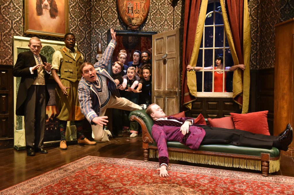 A chaotic scene from The Play That Goes Wrong with a man in a cricket jumper falling into a room, captured in mid-air sailing past another man lying asleep or dead on a chaise longue. Behind them, a group of others cluster round a doorway, staring wide-eyed into the room. 